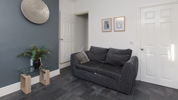 A modern living room with a grey sofa, a glass coffee table, a potted plant, and two framed pictures on the wall.