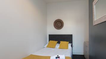 A neatly made bed with a mustard-colored blanket and pillows, a round wall decoration, and a small bedside table with a lamp.