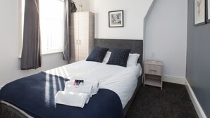 A neatly made bed with white linens and dark blue pillows, a wooden wardrobe, a bedside table with a lamp, and a framed picture on the wall.