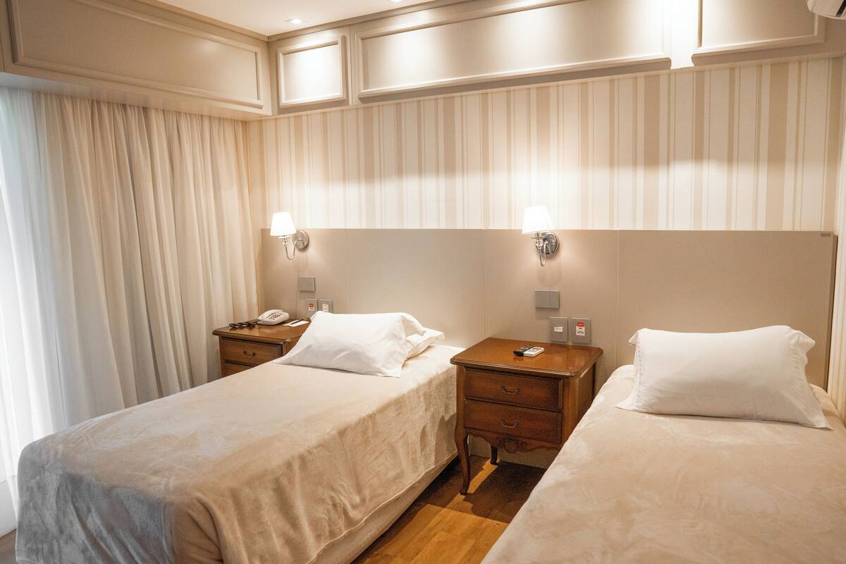 Camera d'albergo con due letti singoli, un comodino in legno con lampada, telefono e un punto luce a parete.