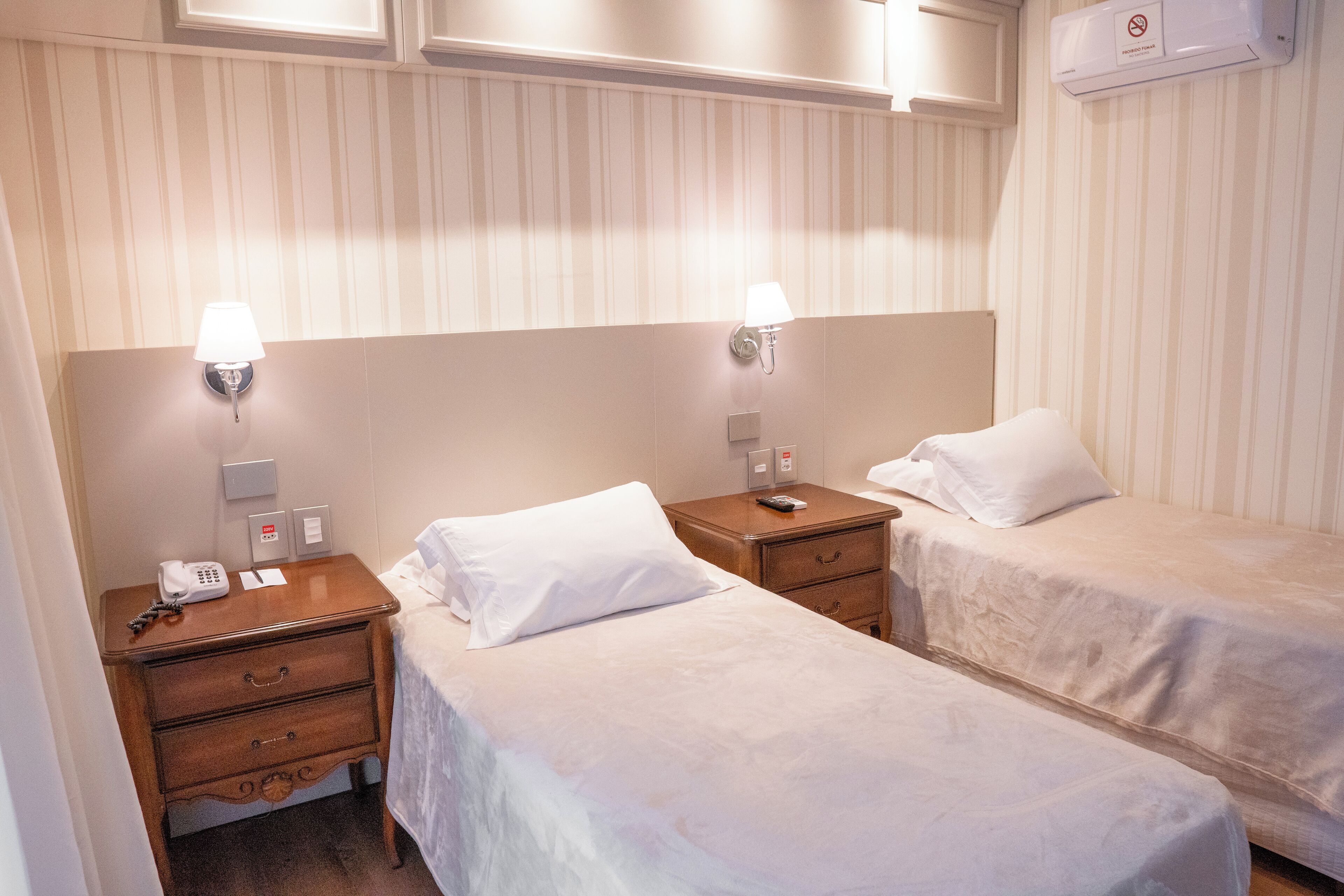 A hotel room with two single beds, a wooden nightstand with a lamp, a telephone, and a wall-mounted air conditioner.