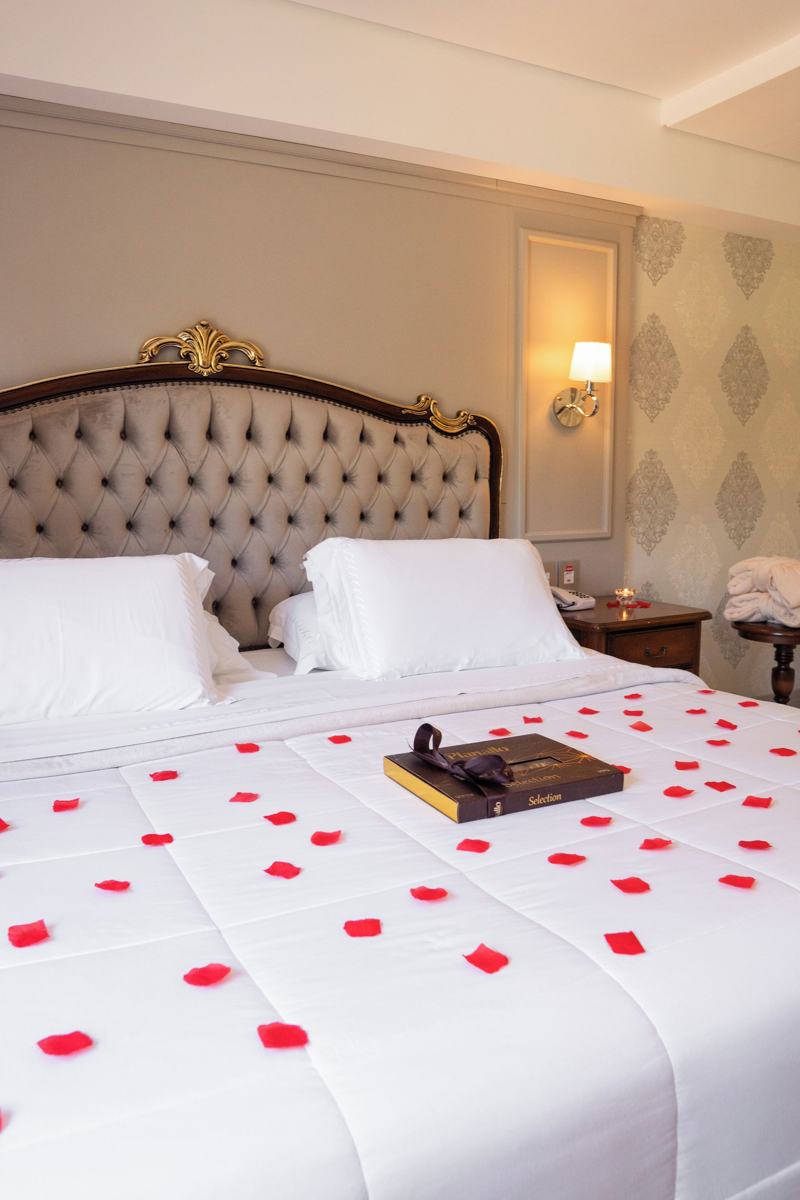 A neatly made bed with white linens and red rose petals, a book, and a bedside lamp.
