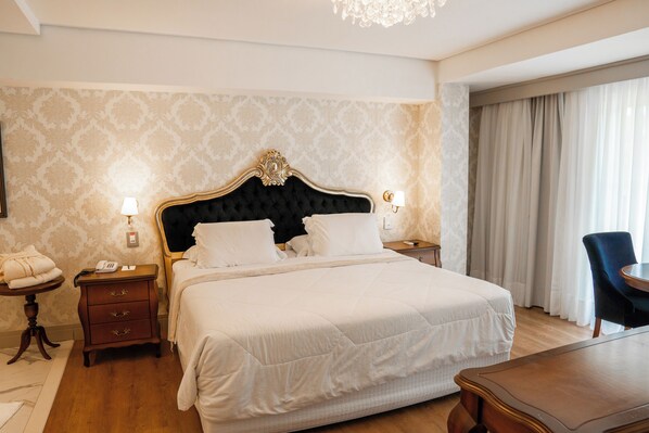 Une chambre d’hôtel avec un grand lit, deux tables de chevet avec des lampes, une petite table en bois, une chaise et un lustre.