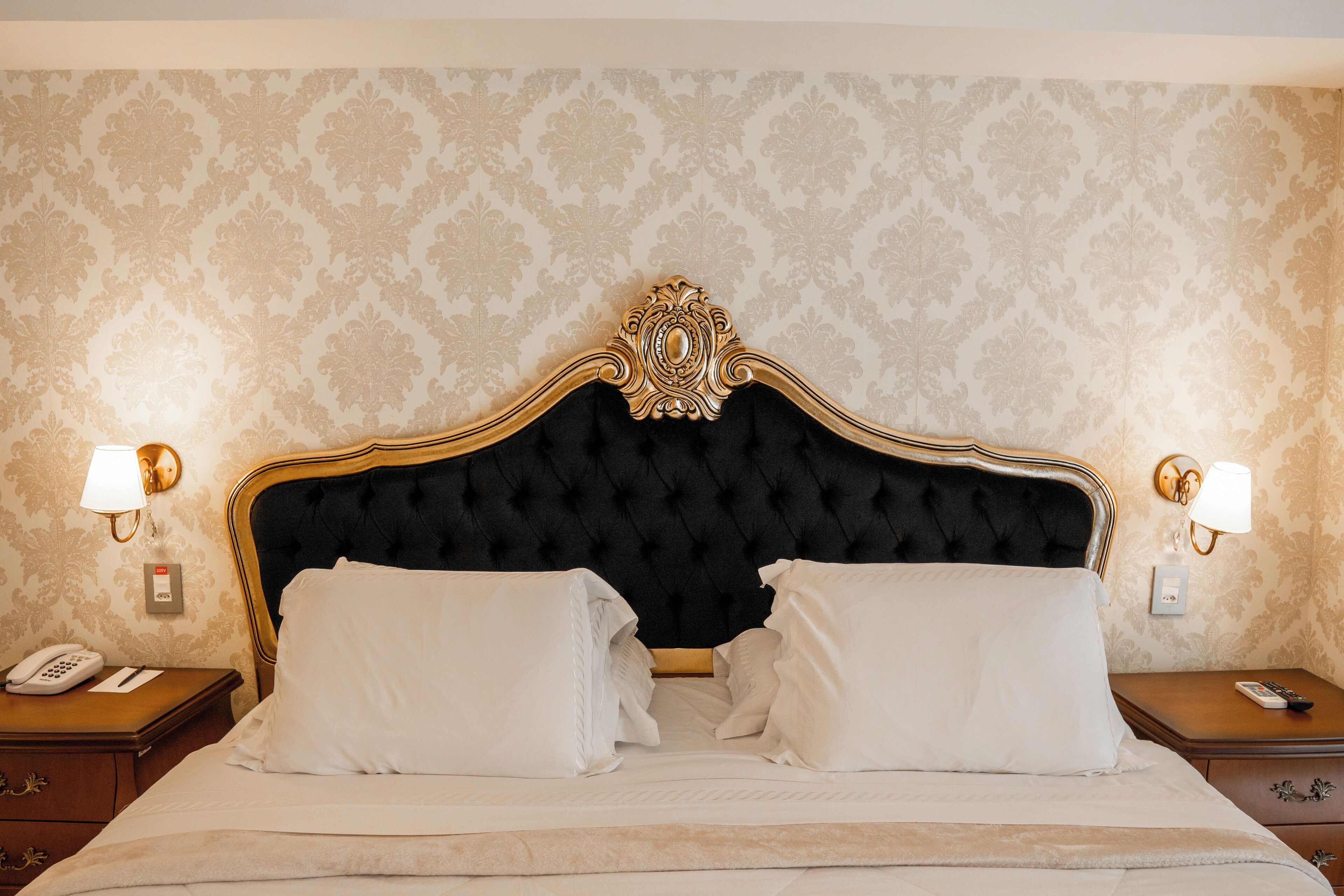 Een hotelkamer met een gestoffeerd hoofdeinde, twee bedlampjes, een telefoon en een netjes opgemaakt bed met kussens.