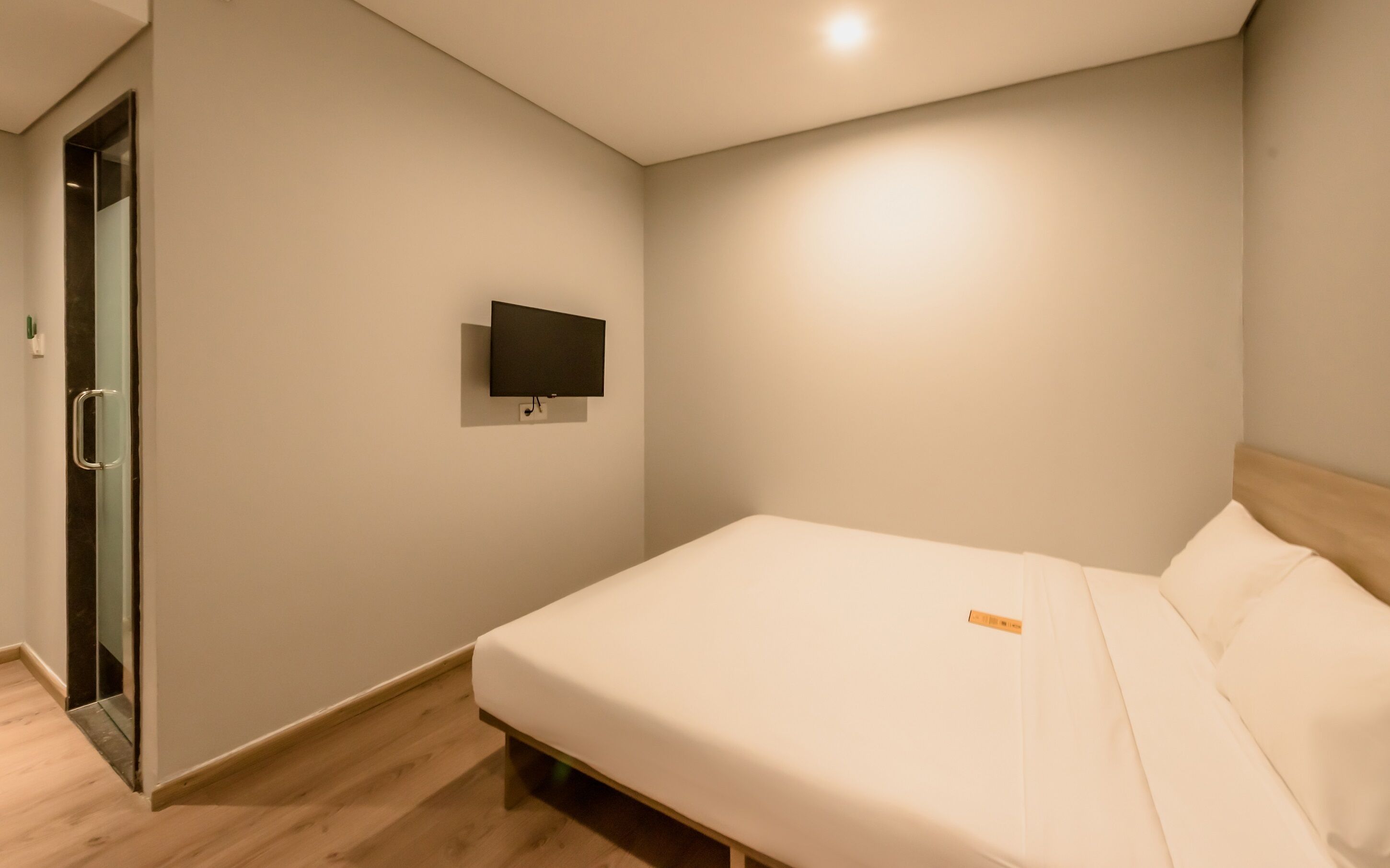 A minimalist bedroom with a single bed, a flat-screen TV mounted on the wall, and a wooden floor.