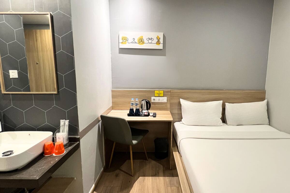 A compact hotel room with a bed, a desk with a chair, a sink, and a mirror.