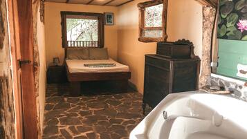 Un dormitorio rústico con suelo de piedra, cama de madera, cómoda y ventana con vistas a árboles.