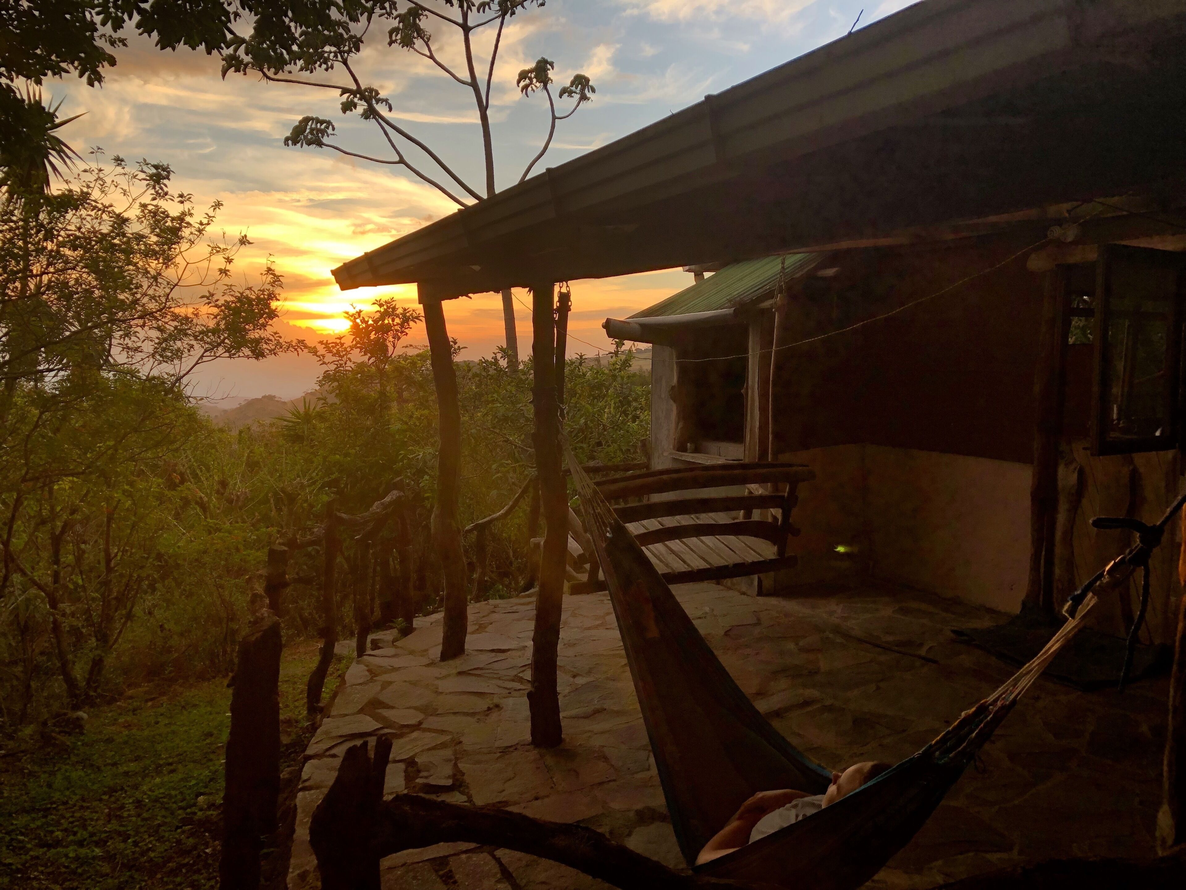 Una terraza de madera con una hamaca, un camino de piedra y un edificio con techo verde.