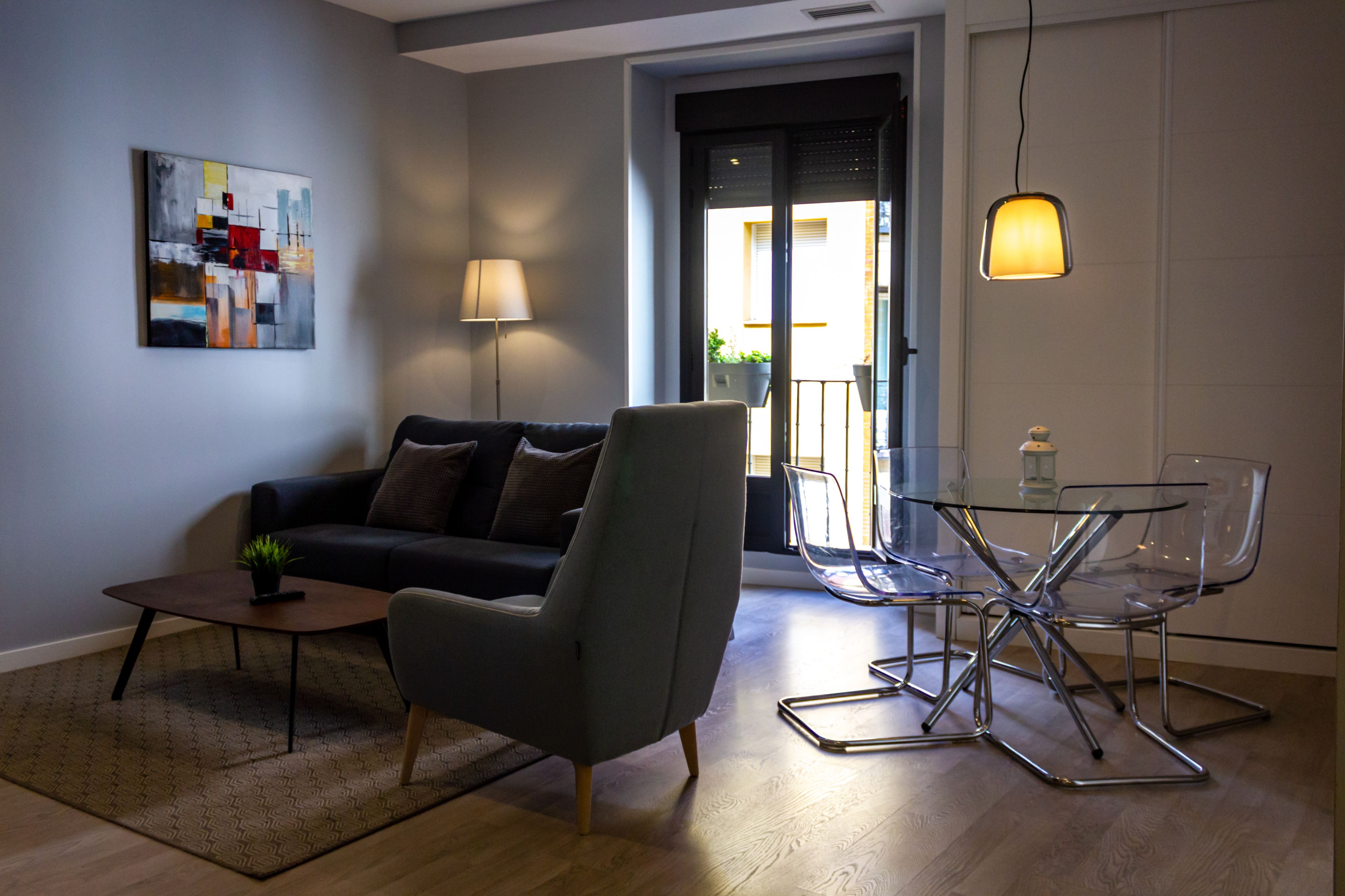 A modern living room with a grey sofa, a small coffee table, and a glass dining table with chairs. There is a painting on the wall and a lamp on the table.