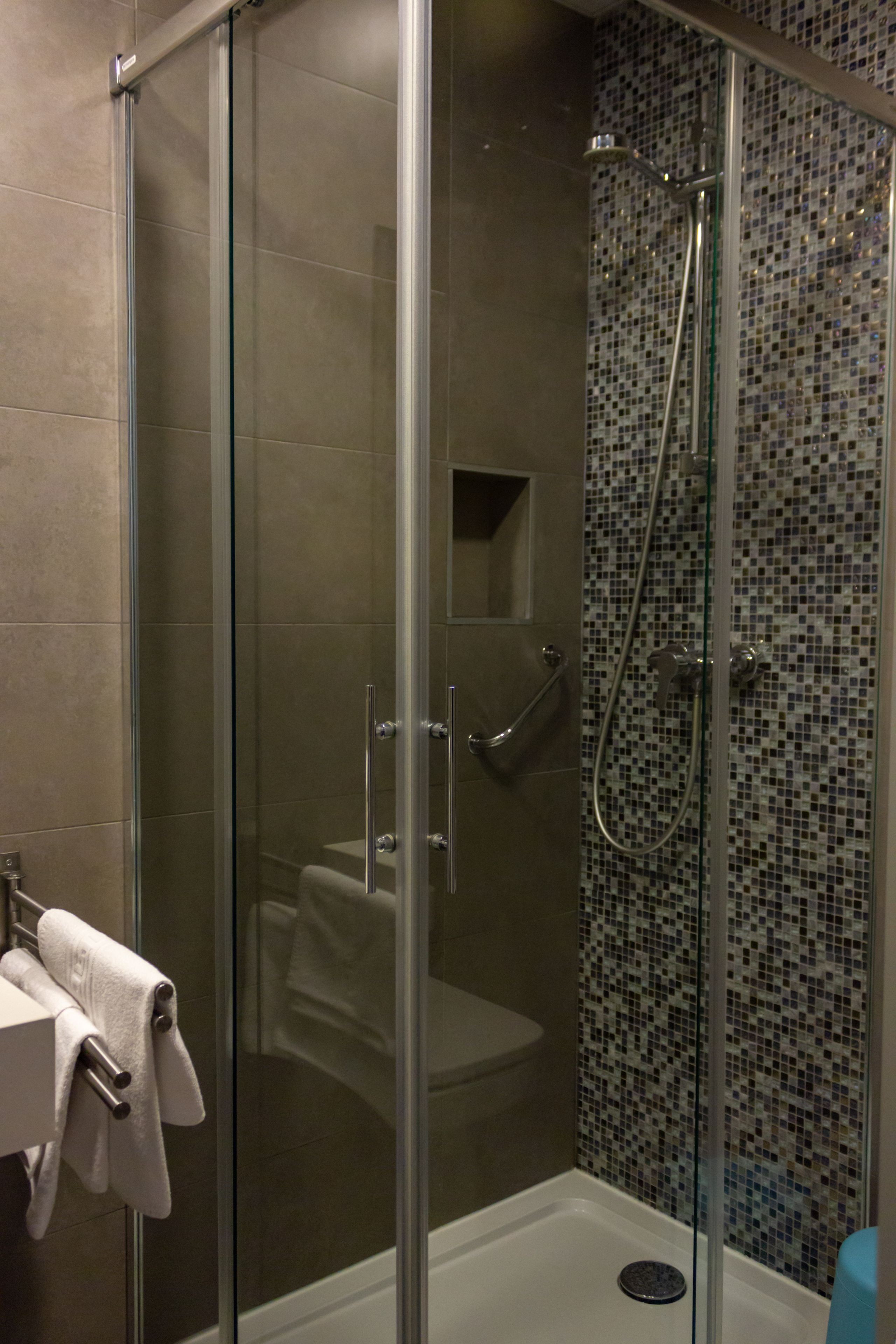 A glass-enclosed shower with a tiled mosaic wall, a visible toilet, and a towel rack with towels.