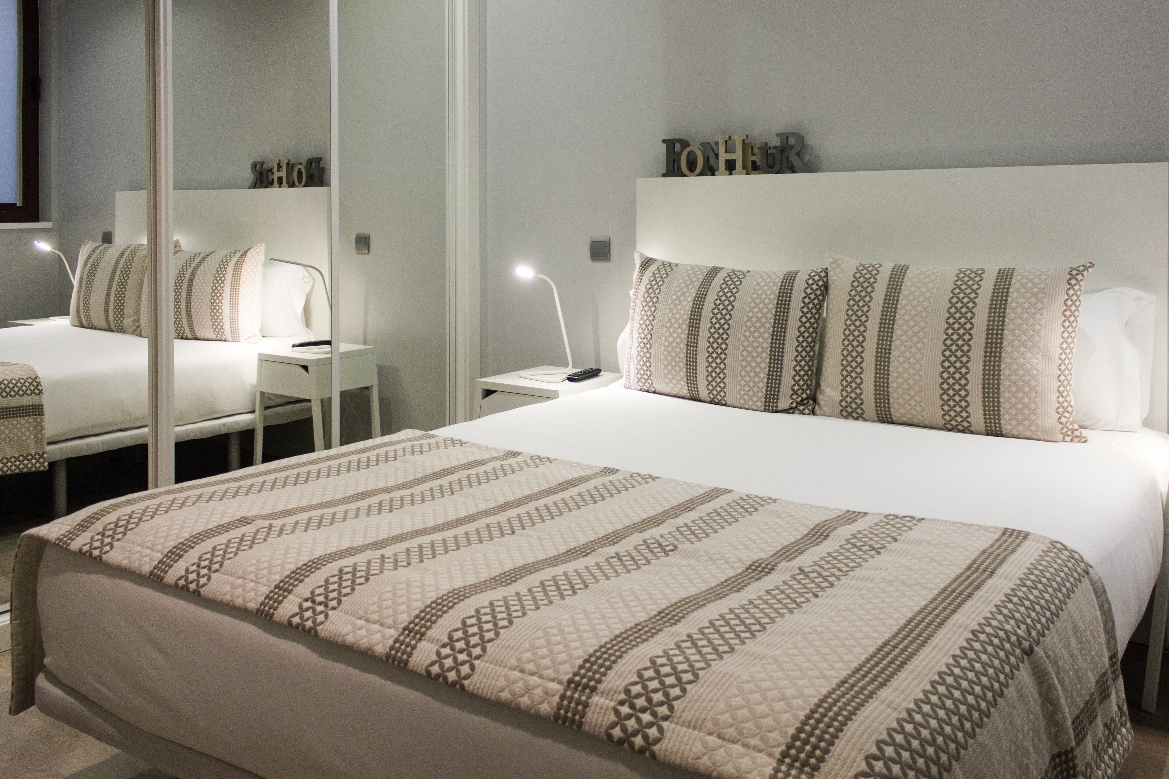 Quarto de hotel com uma cama grande, mesa de cabeceira com abajur, espelho e cabeceira com letras decorativas.