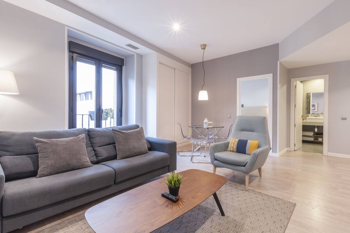A modern living room with a grey sofa, a wooden coffee table, and a glass dining table with chairs.