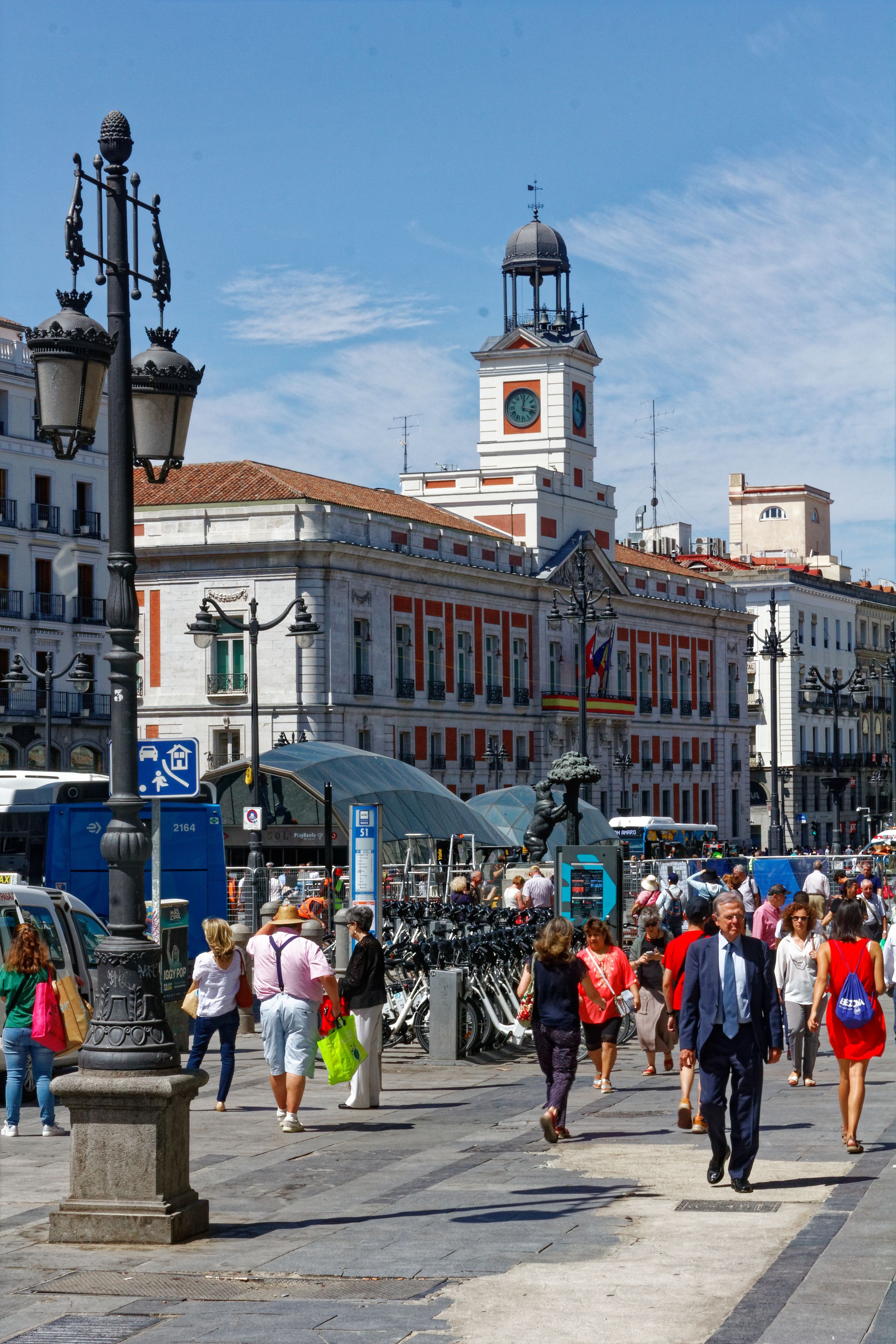 A busy pedestrian area with historic buildings, a clock tower, and a clear blue sky.