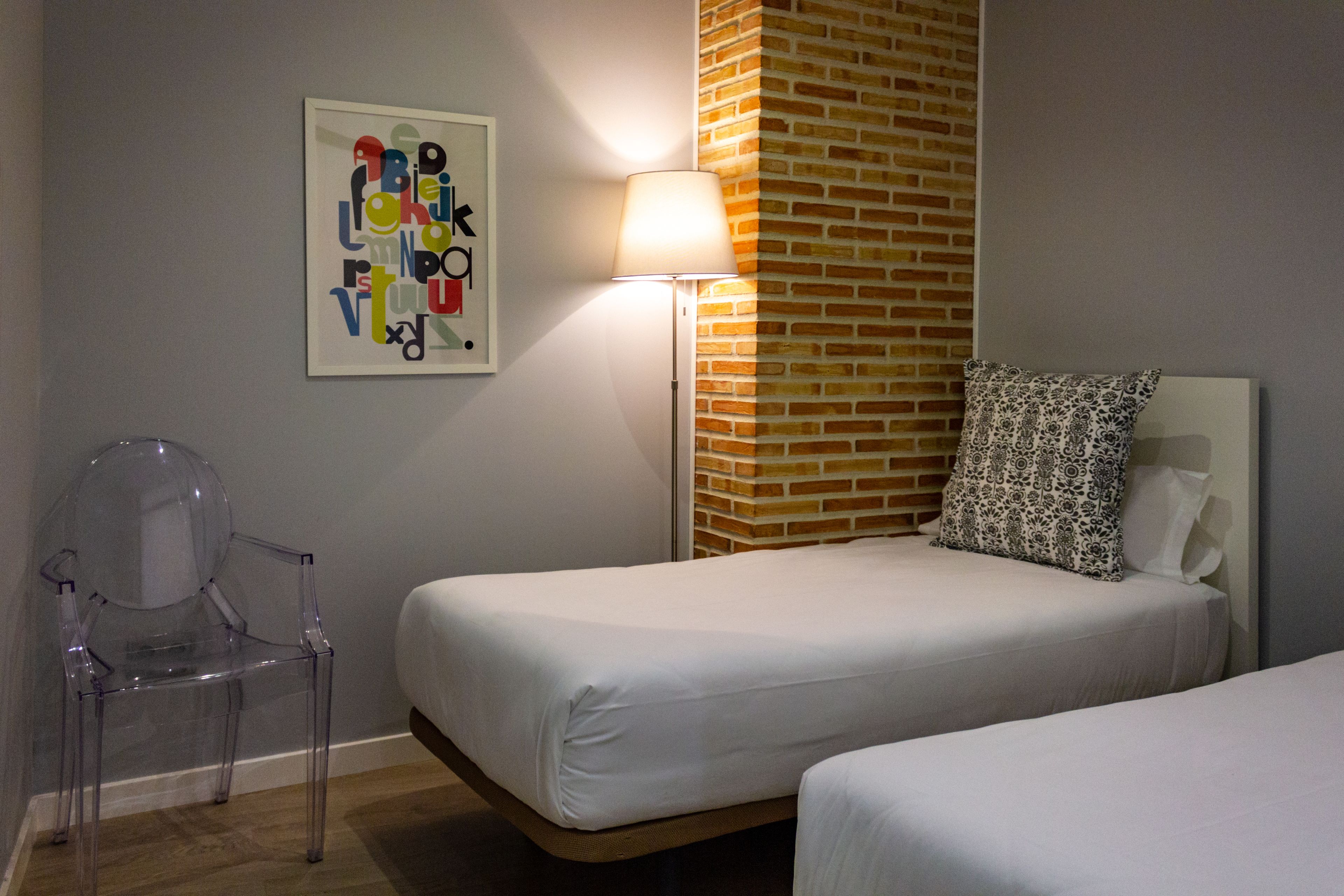 Quarto de hotel com duas camas, uma cadeira de plástico transparente, um abajur e uma parede de tijolos.