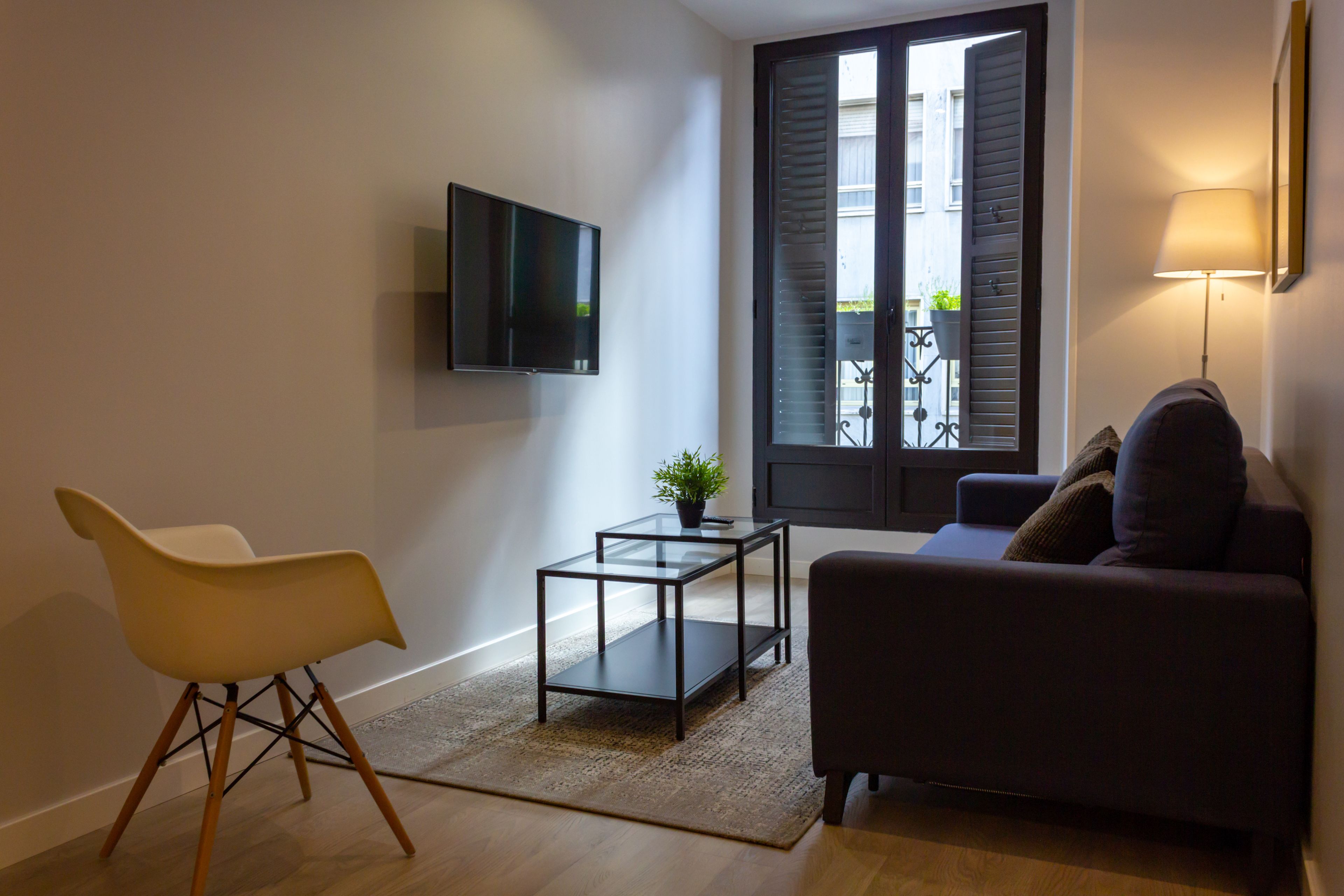 Una sala de estar moderna con un sofá, una mesa de centro de vidrio, un televisor montado en la pared y un ventanal con persianas.