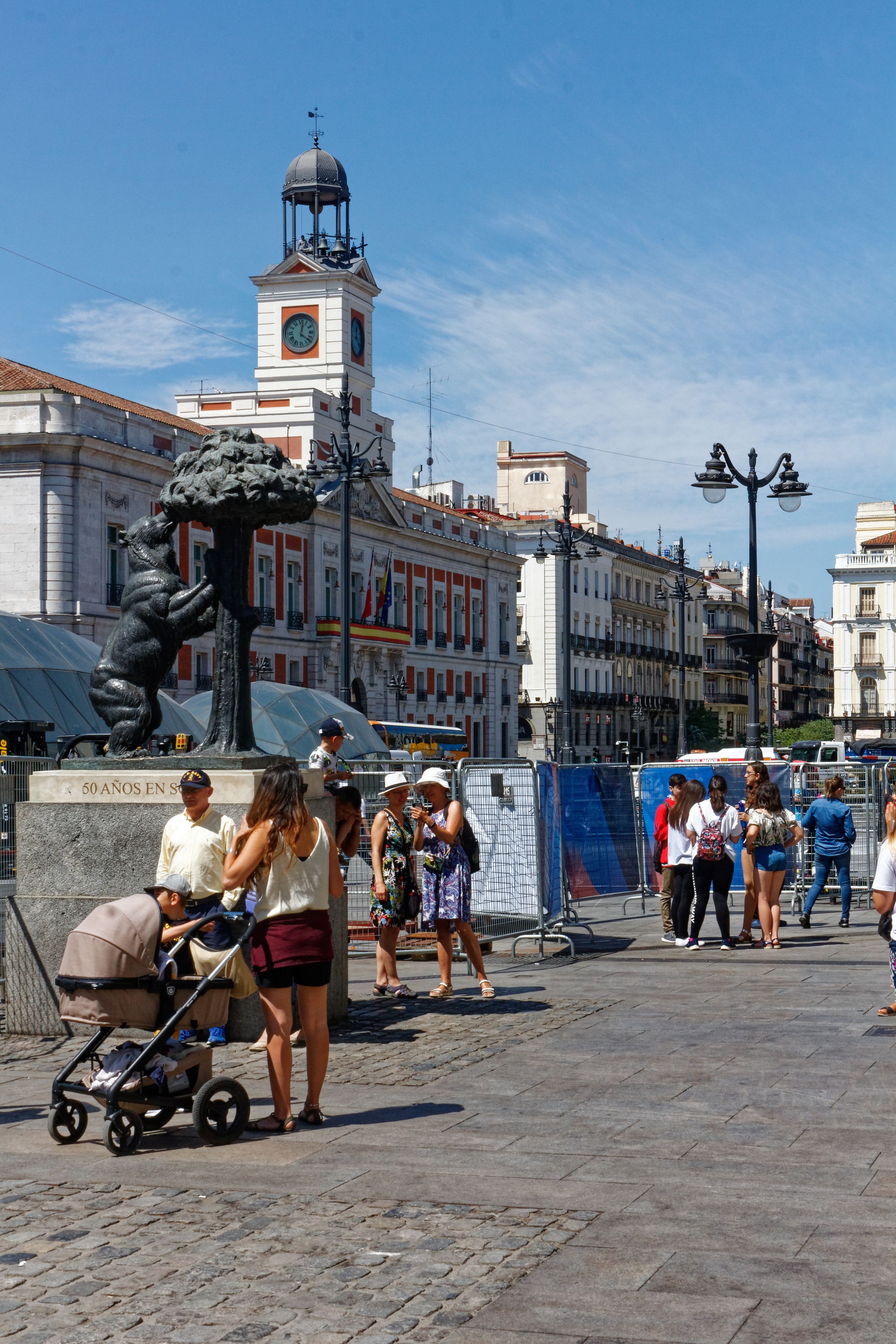 A public square with a statue, a clock tower, and people walking.