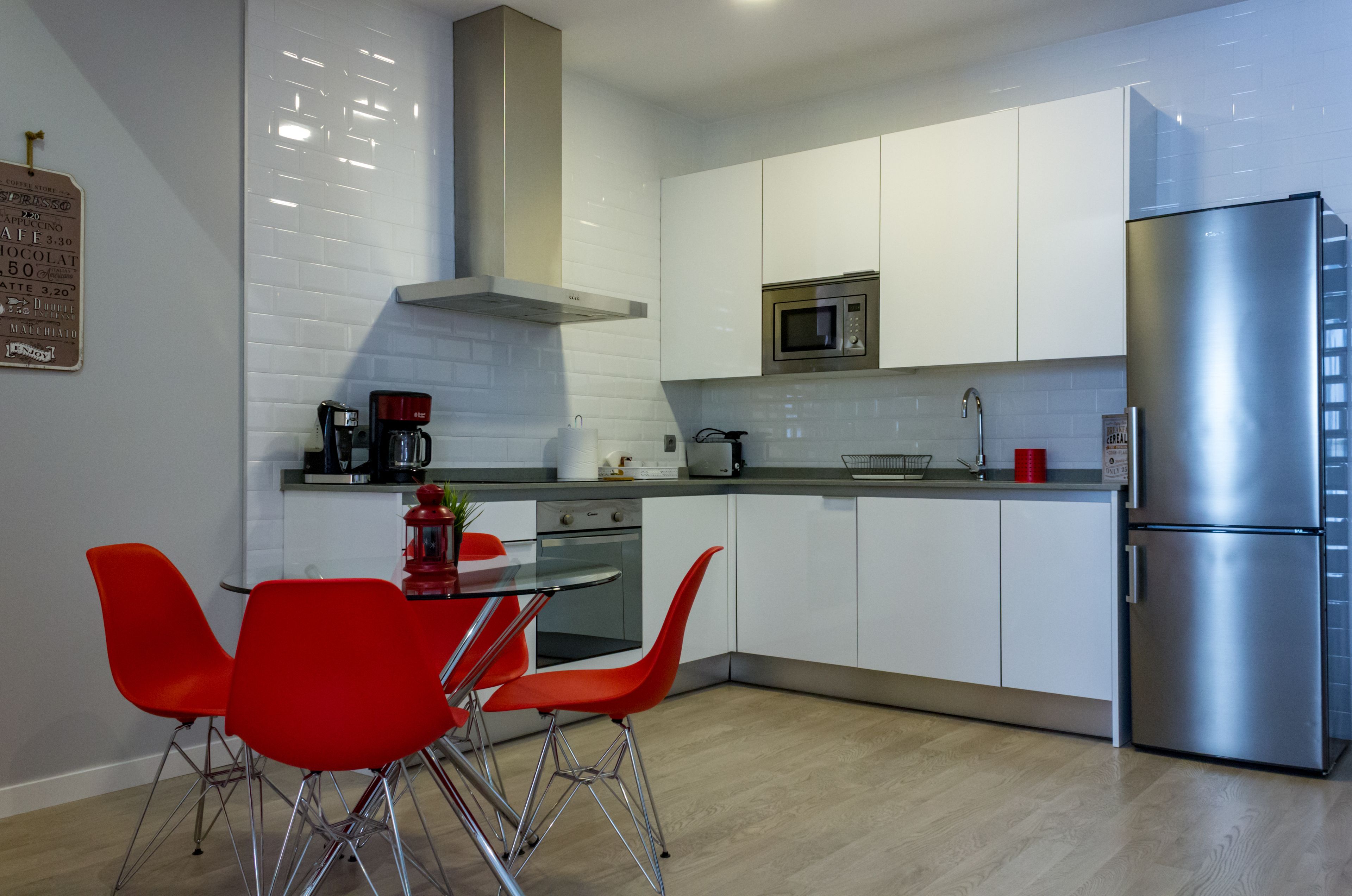 A modern kitchen with white cabinets, a stainless steel refrigerator, a microwave, and a coffee maker. There is a dining table with red chairs.