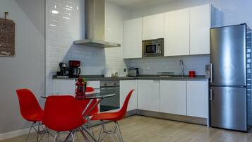 A modern kitchen with white cabinets, a stainless steel refrigerator, a microwave, and a coffee maker. There is a dining table with red chairs.