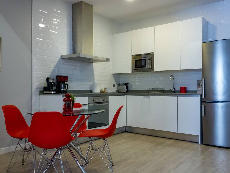 A modern kitchen with white cabinets, a stainless steel refrigerator, a microwave, and a coffee maker. There is a dining table with red chairs.