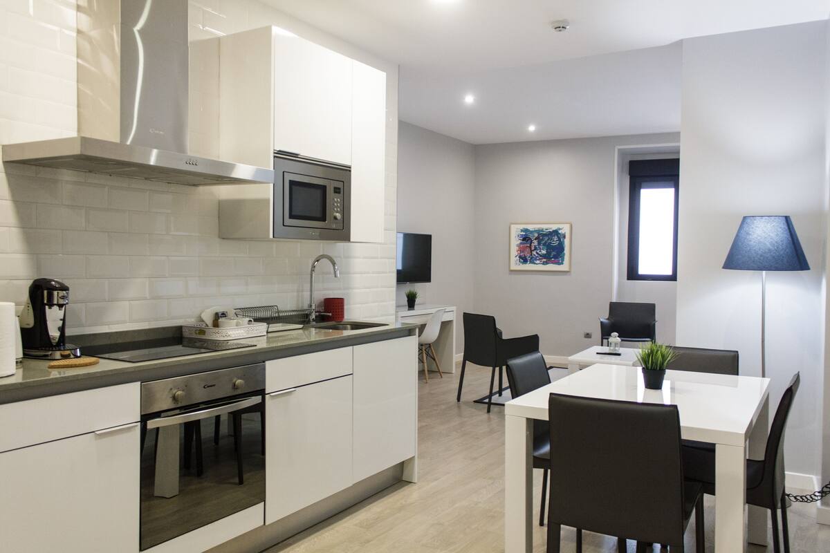 A modern kitchen with white cabinetry, a built-in microwave, and a coffee maker. There is a dining area with a white table and black chairs.