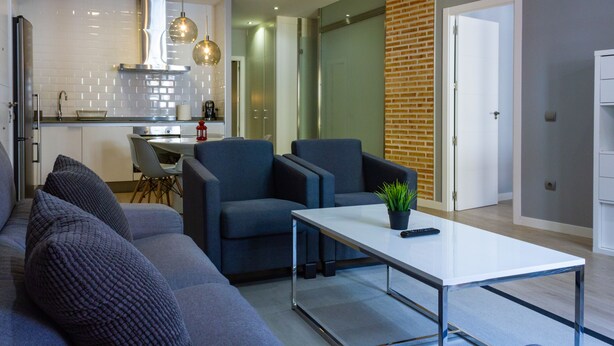 A modern living room with a grey sofa, two armchairs, and a coffee table. There is a kitchen area with a stove, sink, and hanging pendant lights. - Arenal Suites (Madrid)