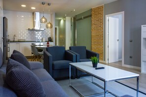 A modern living room with a grey sofa, two armchairs, and a coffee table. There is a kitchen area with a stove, sink, and hanging pendant lights. - Arenal Suites (Madrid)