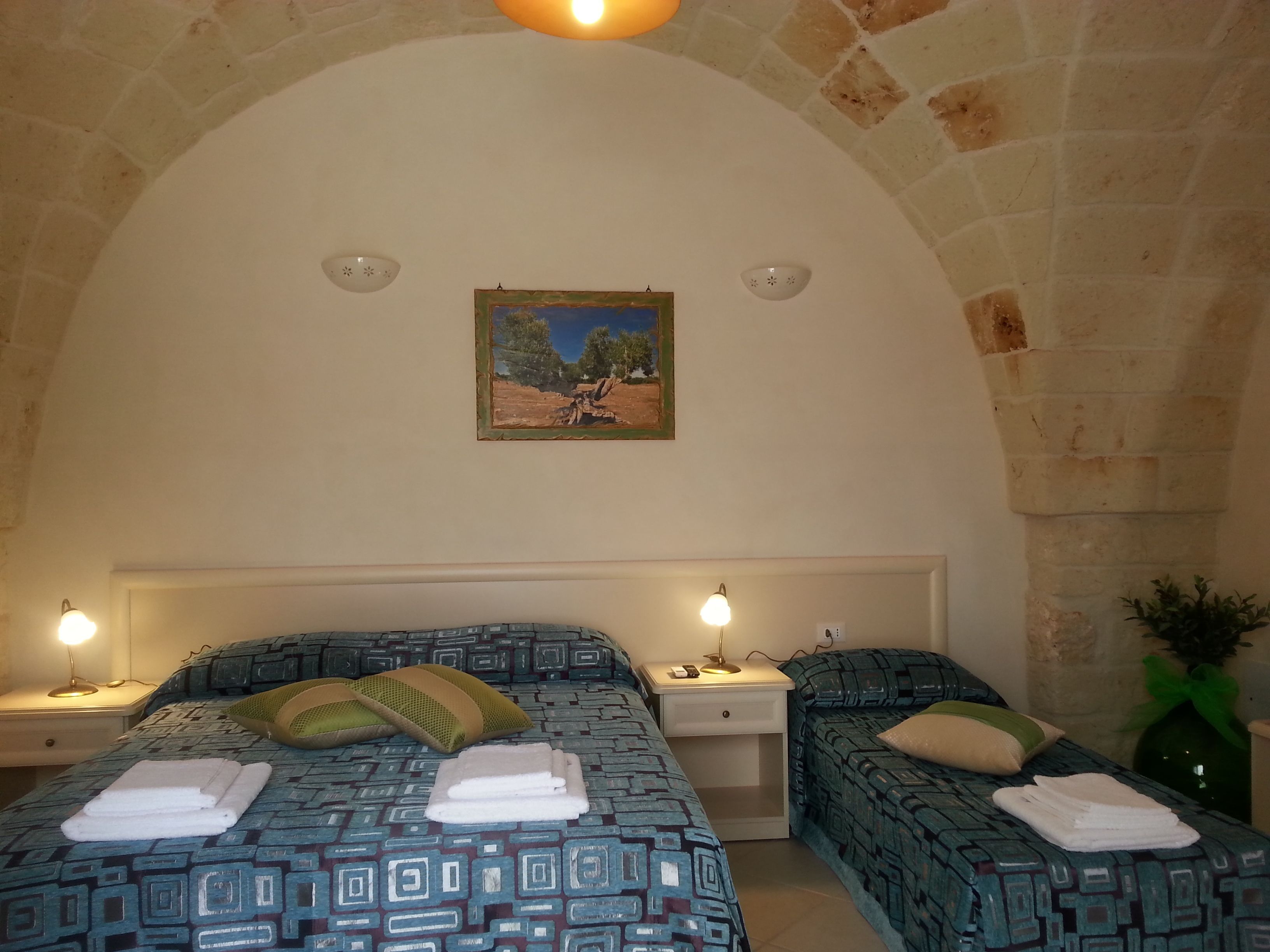 A room with two beds, each with a patterned bedspread and pillows, a nightstand with a lamp, and a painting on the wall.