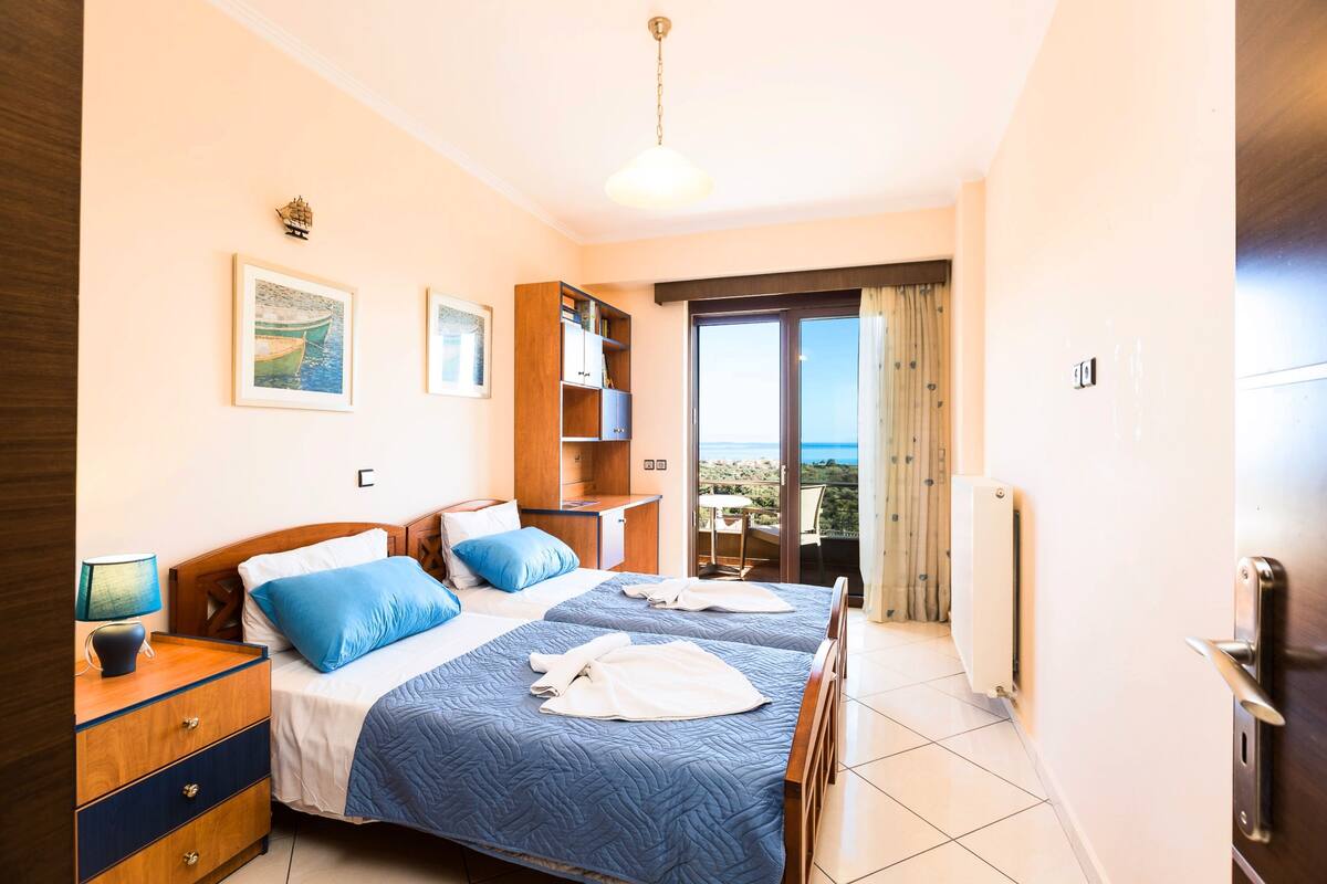 A hotel room with two beds, a wooden dresser, a nightstand, a bookshelf, and a view of the outdoors.
