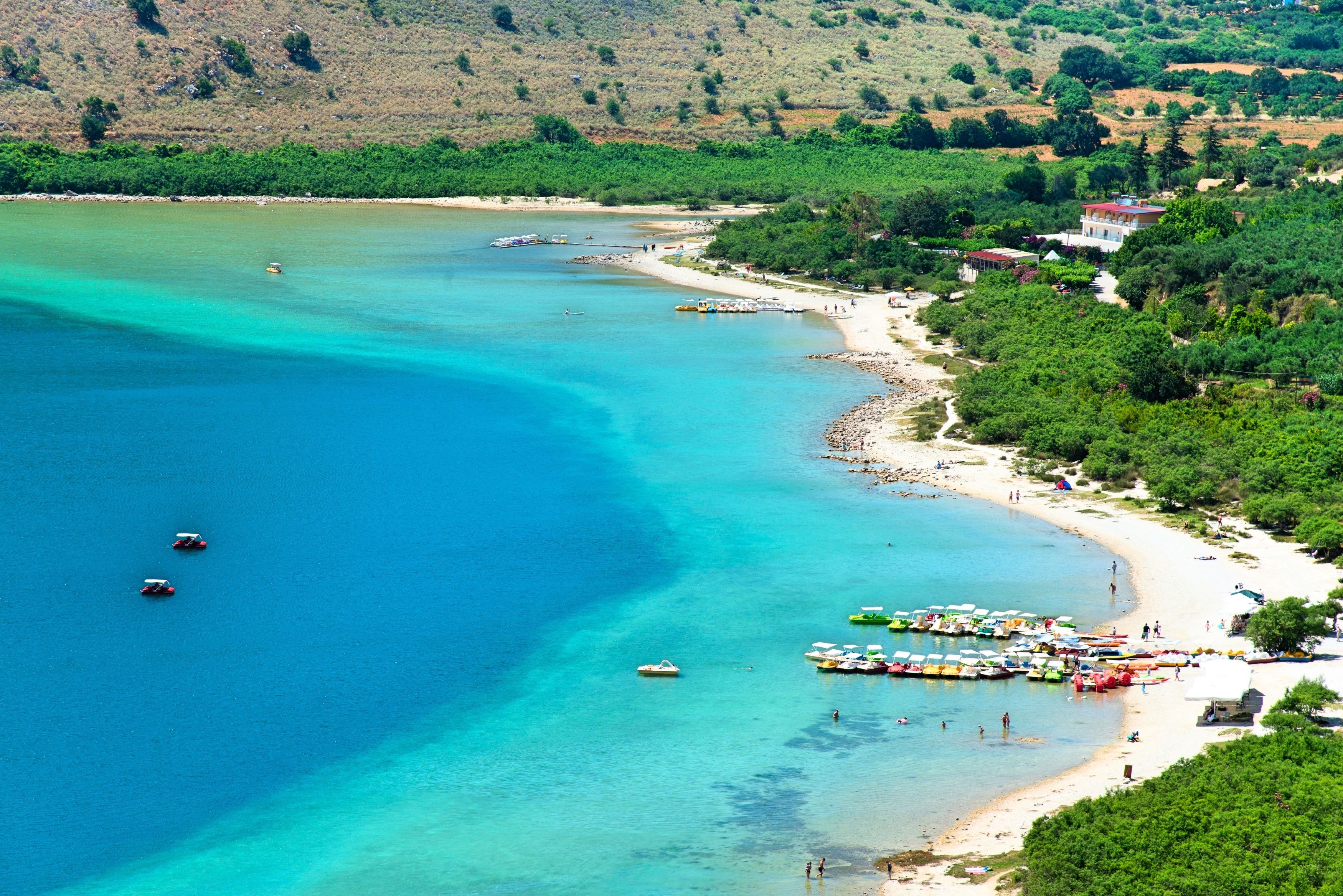 A coastal area with clear blue waters, boats, and a beach with umbrellas.