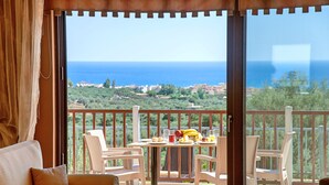 Un balcón con mobiliario de mimbre, una mesa con fruta y vasos, y vistas al mar y vegetación.