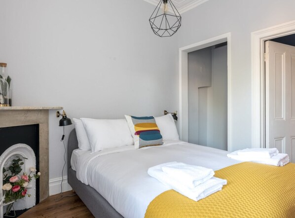 A bedroom with a bed, a bedside table, a lamp, a fireplace, and a door.