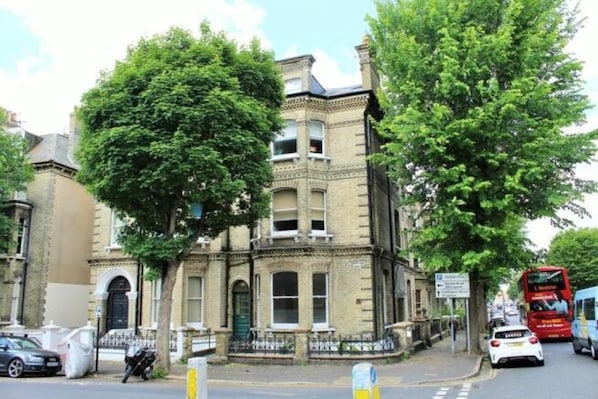 A historic building with a prominent corner entrance, a large tree in front, and a red double-decker bus on the street.