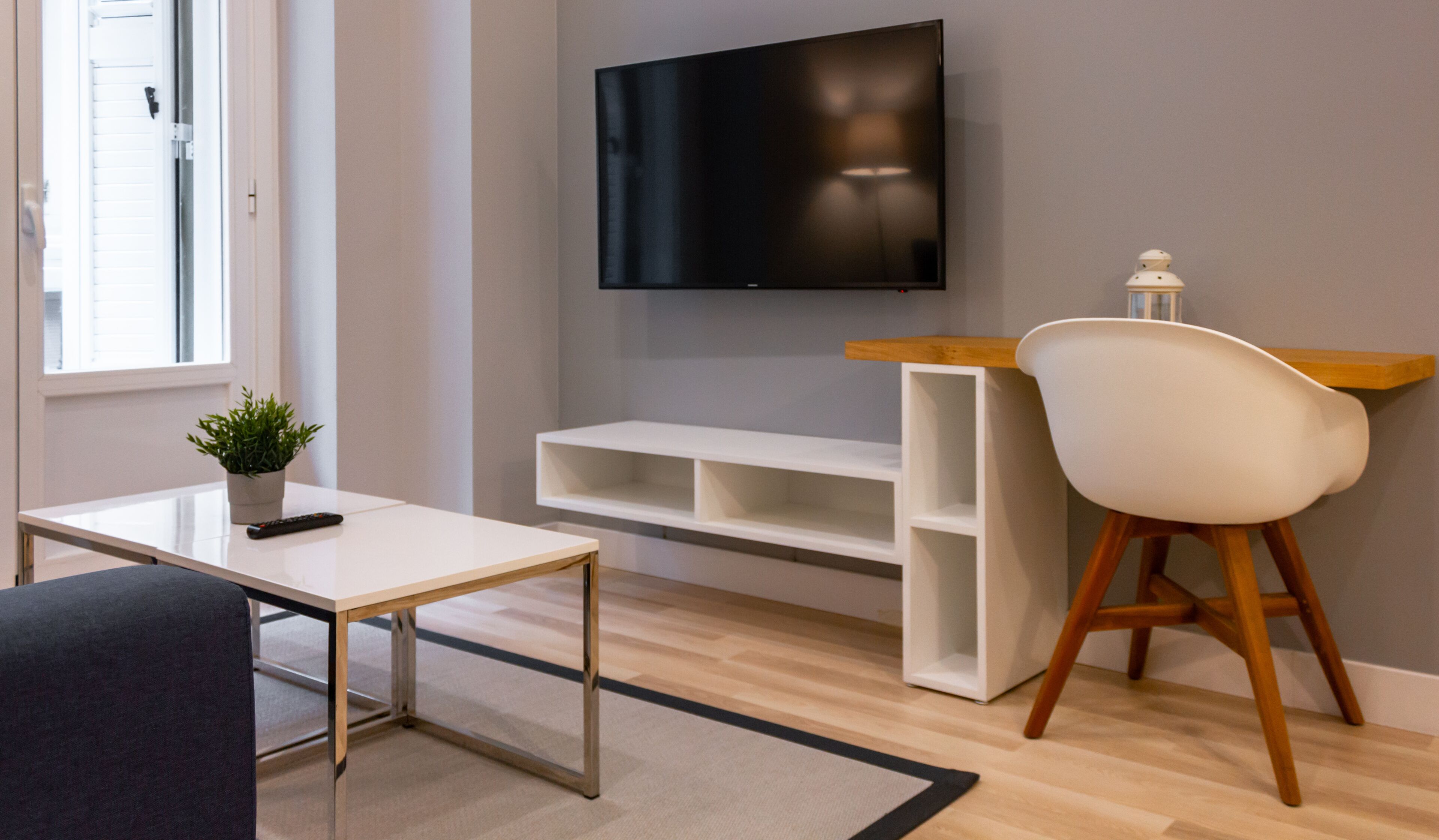A modern living room with a flat-screen TV mounted on the wall, a white coffee table, a dark sofa, a wooden side table, and a white chair.