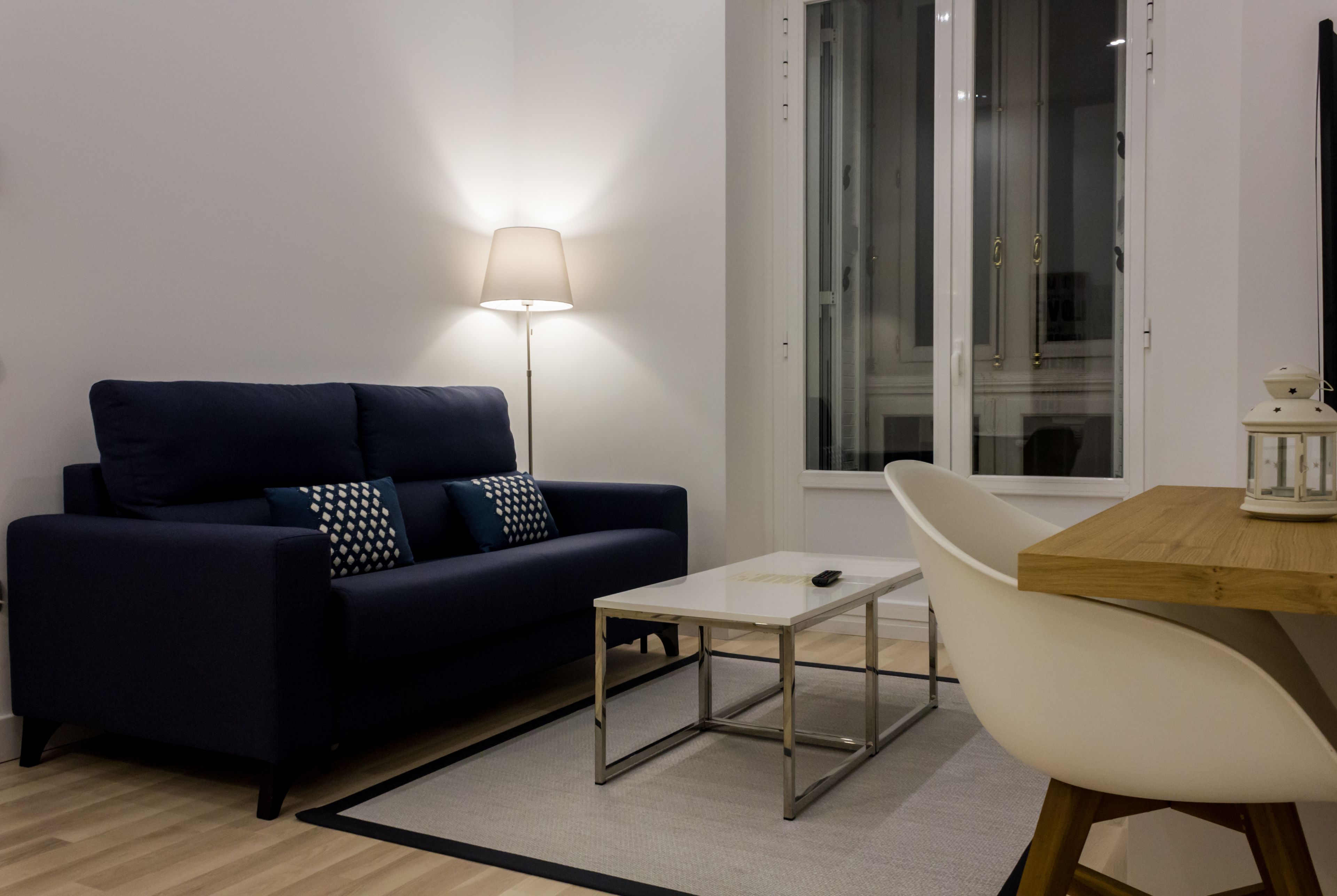 A modern living room with a dark blue sofa, a small white table, a white chair, and a floor lamp.