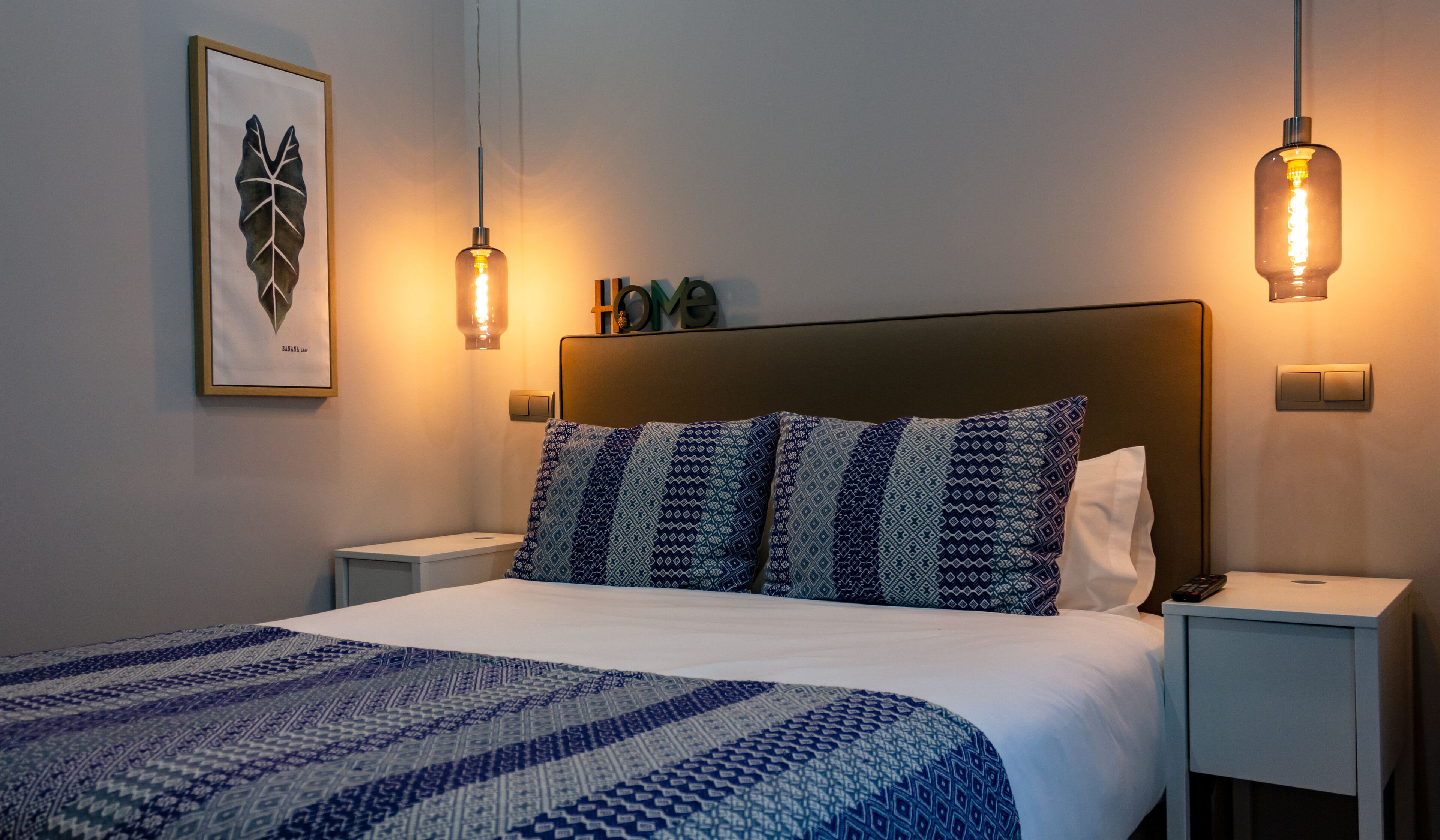 Een keurig opgemaakt bed met beddengoed met een patroon, twee nachtkastjes met lampen, een lijst met kunstwerk aan de muur, en een ‘HOME’ bord boven het bed.