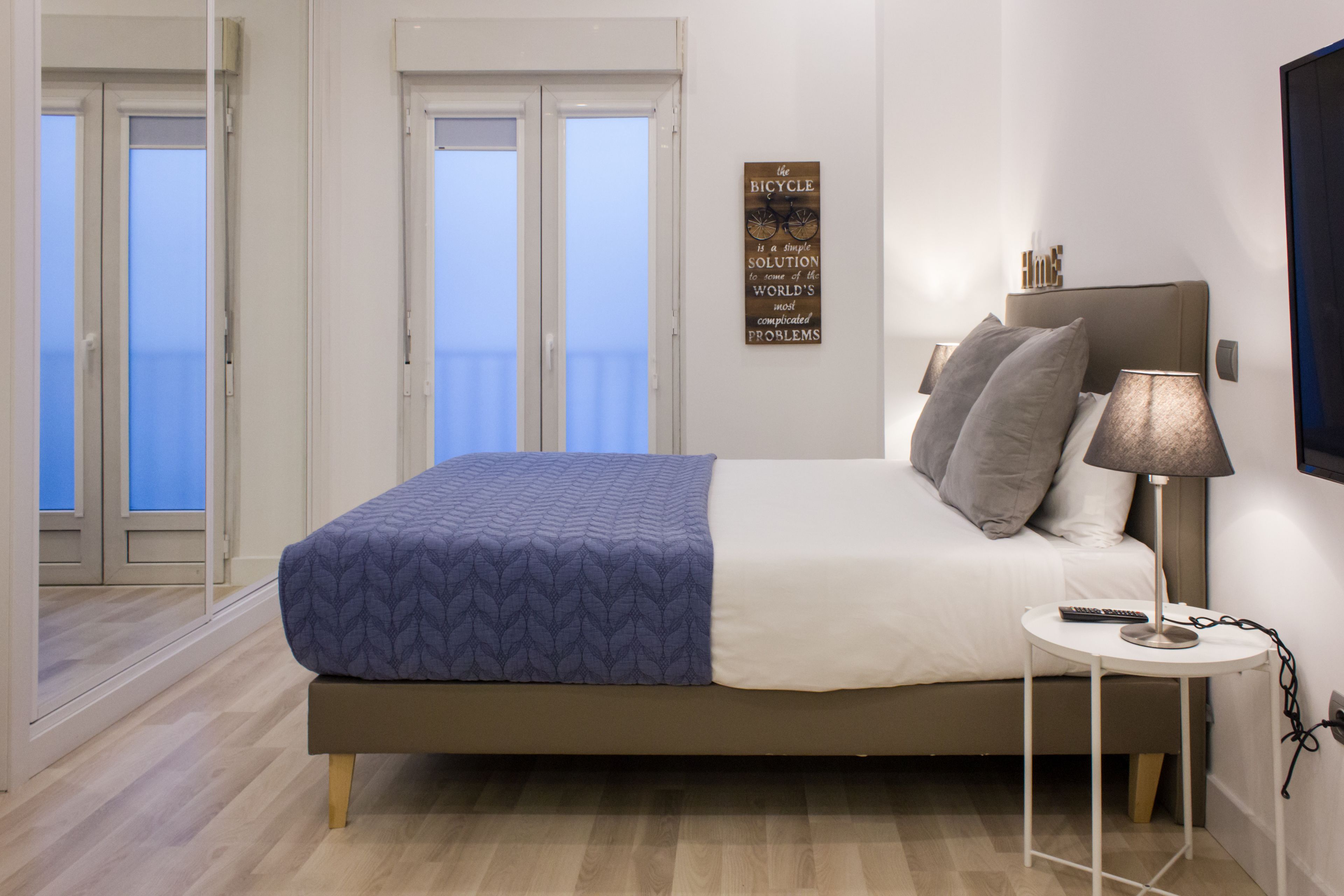 A bedroom with a bed, a bedside table with a lamp, a wall-mounted TV, and a door leading to a balcony.