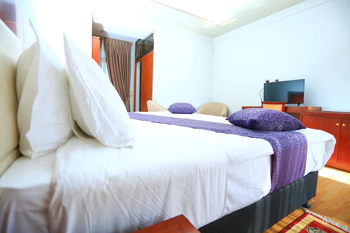 A neatly made bed with white and purple bedding, a television on a wooden stand, and a wooden floor.