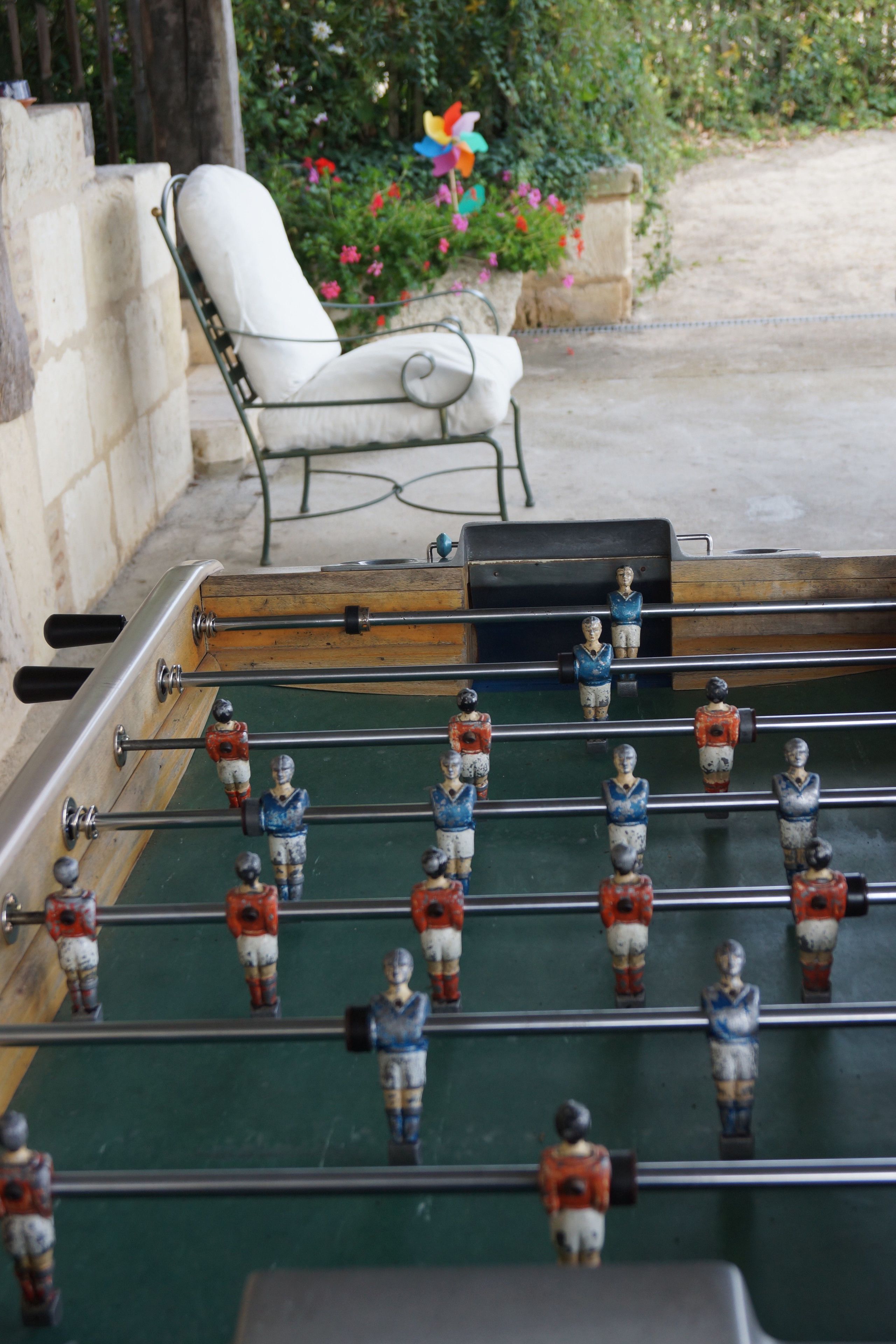 A foosball table with miniature figures in red and blue uniforms, positioned in a room with a stone wall and a white chair.