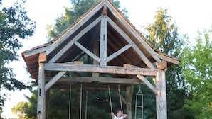 A wooden swing set with a triangular roof structure, supported by sturdy posts, in a grassy outdoor area with trees in the background. - Domaine de la Mouthe (Saint-Remy)