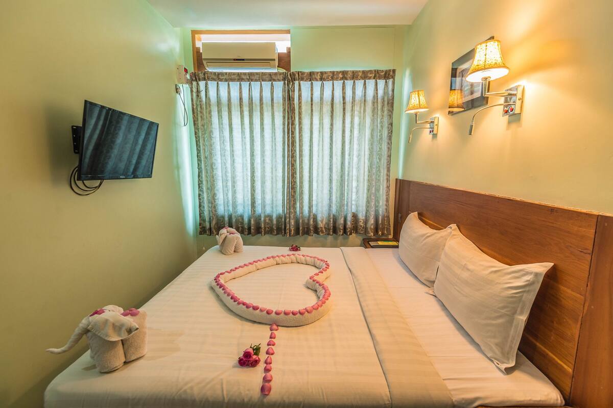 A hotel room with a bed, a television mounted on the wall, a window with curtains, and wall-mounted lights.