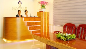 Un comptoir d’accueil d’hôtel avec deux employés, un espace d’accueil en bois, une table à manger avec des chaises et un pot de fleurs.