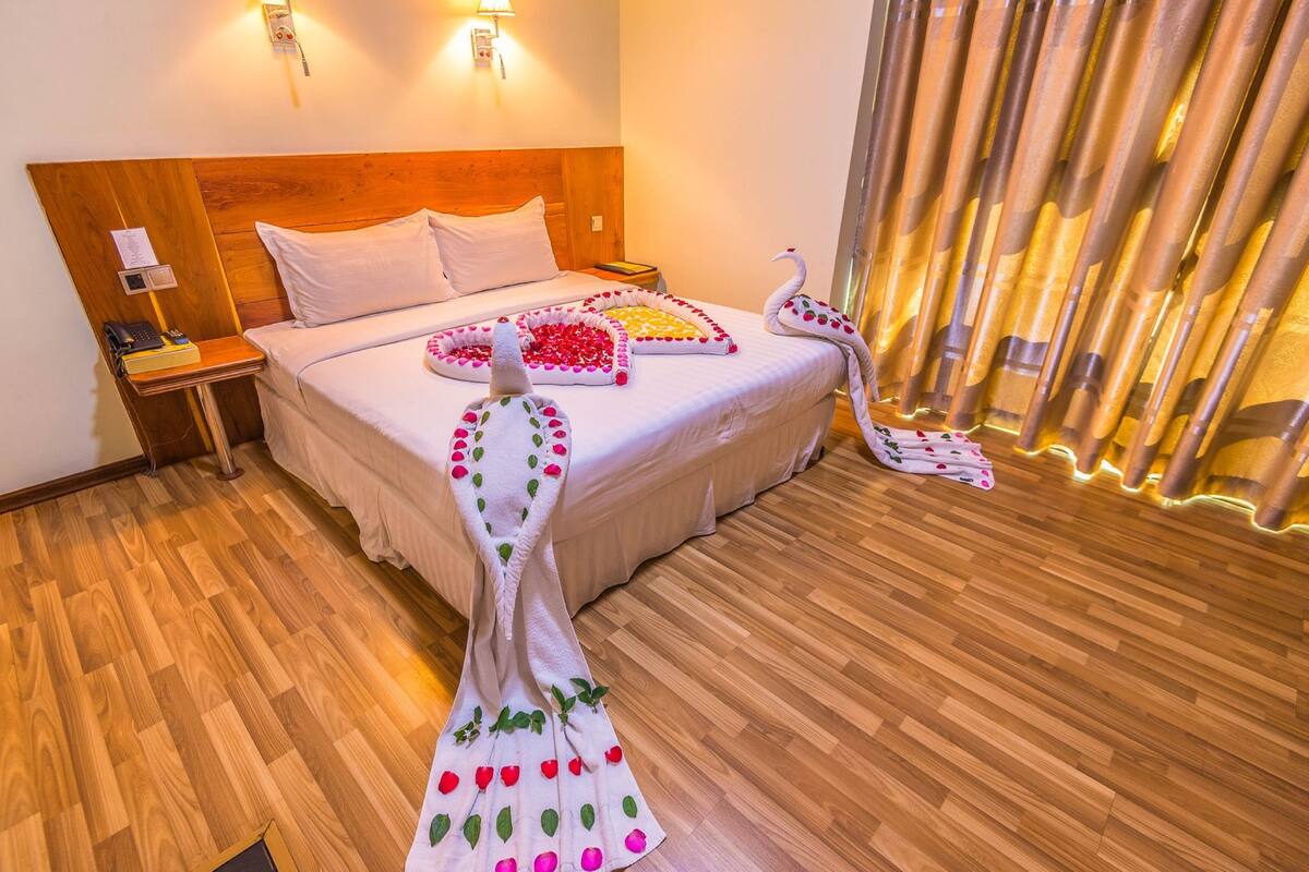 A hotel room with a wooden headboard, a neatly made bed with white linens, a bedside table with a lamp, and a decorative swan made of flowers on the bed.