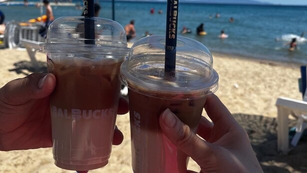 Two iced drinks with straws on a beach with umbrellas and people in the background. - Sevo Hotel (Ayvalik)