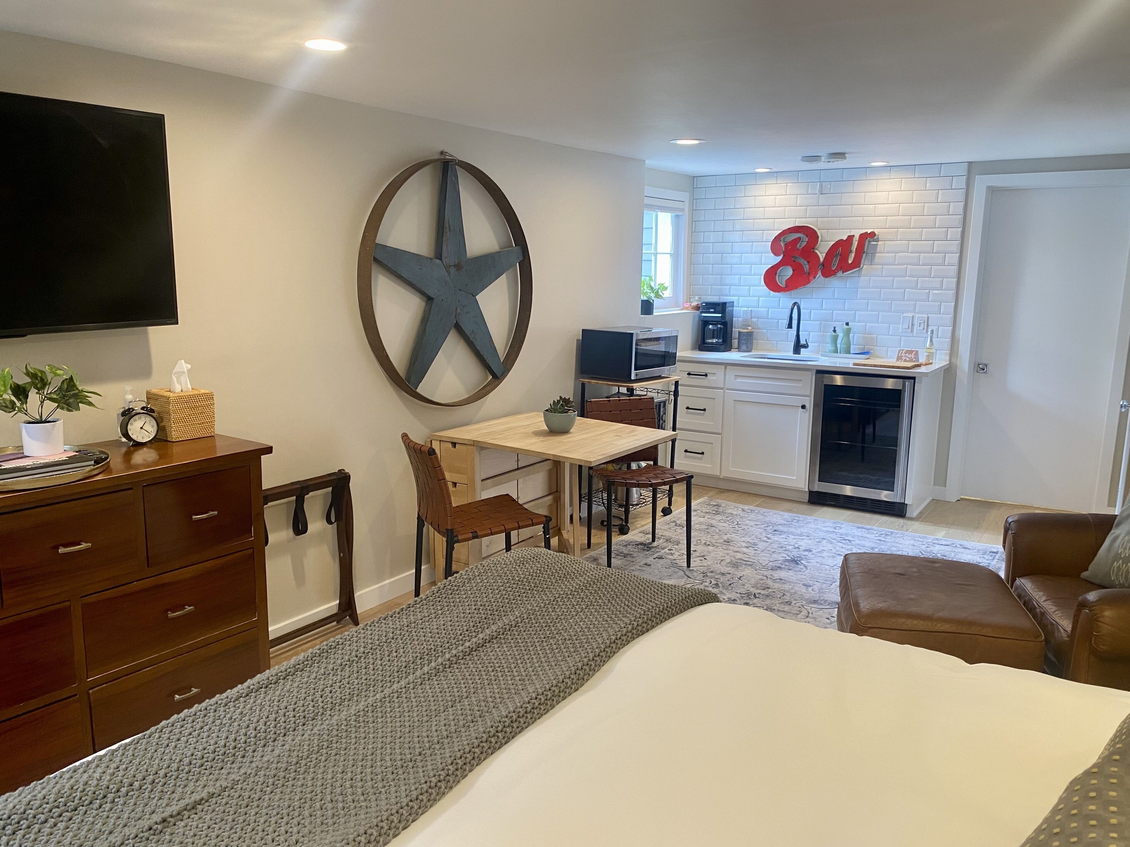 Ein kompaktes Hotelzimmer mit Bett, Schreibtisch, Stuhl, Fernseher, einer großen Wanddekoration, einer Küchenzeile mit einem 'Bar'-Schild und einem braunen Ledersofa.