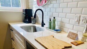 Une cuisine avec un plan de travail blanc, un évier, une machine à café et une planche à découper.
