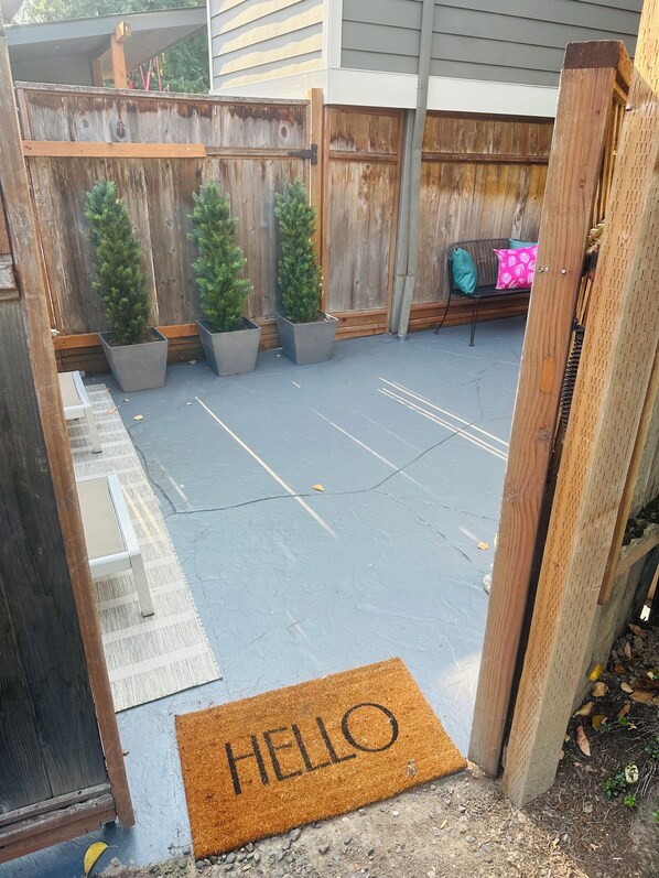 Un patio trasero con valla de madera, tres abetos navideños en macetas, una banca con cojín rosa y un felpudo que dice 'HELLO'.