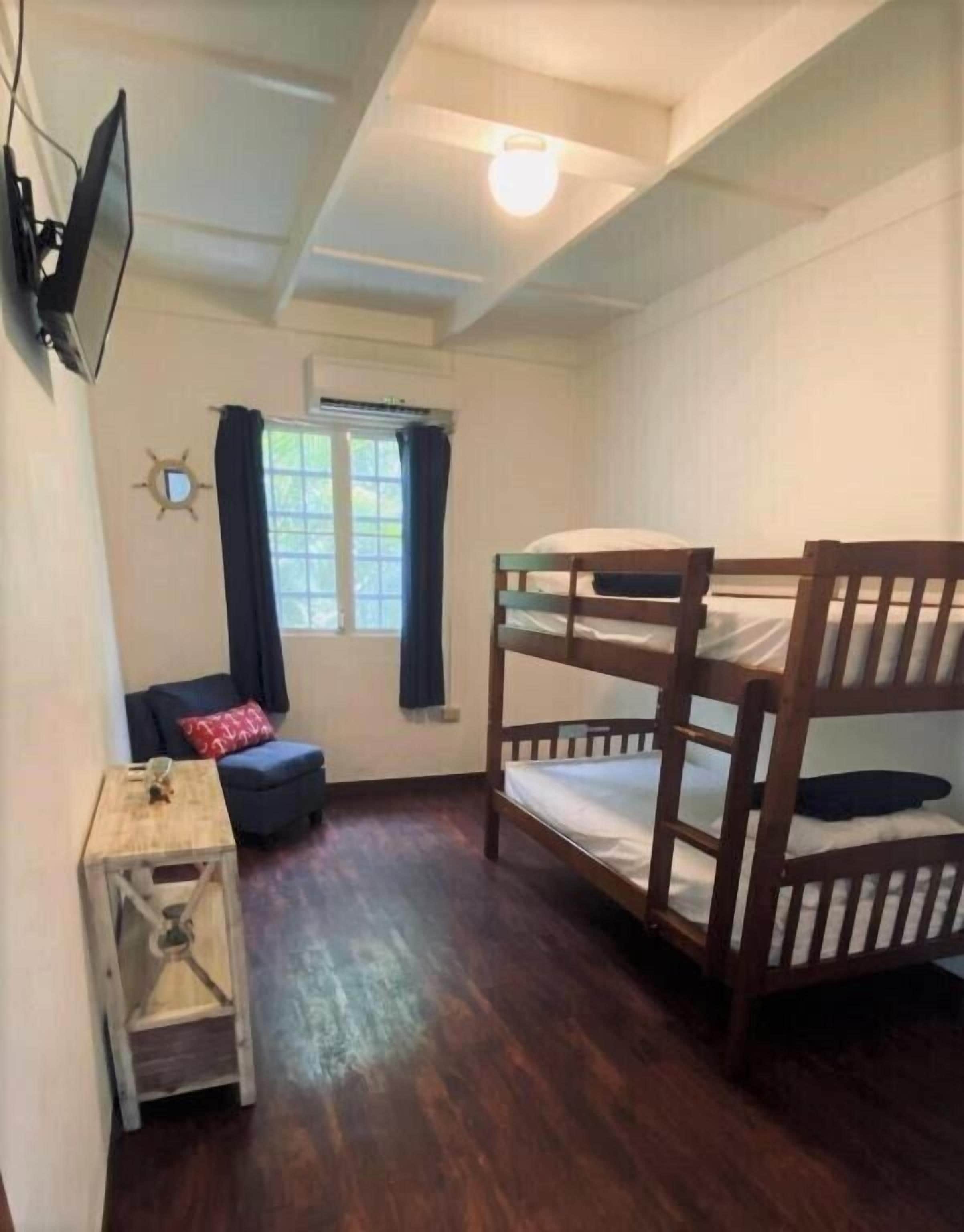 A room with a bunk bed, a small table with a lamp, a window with curtains, and a TV mounted on the wall.