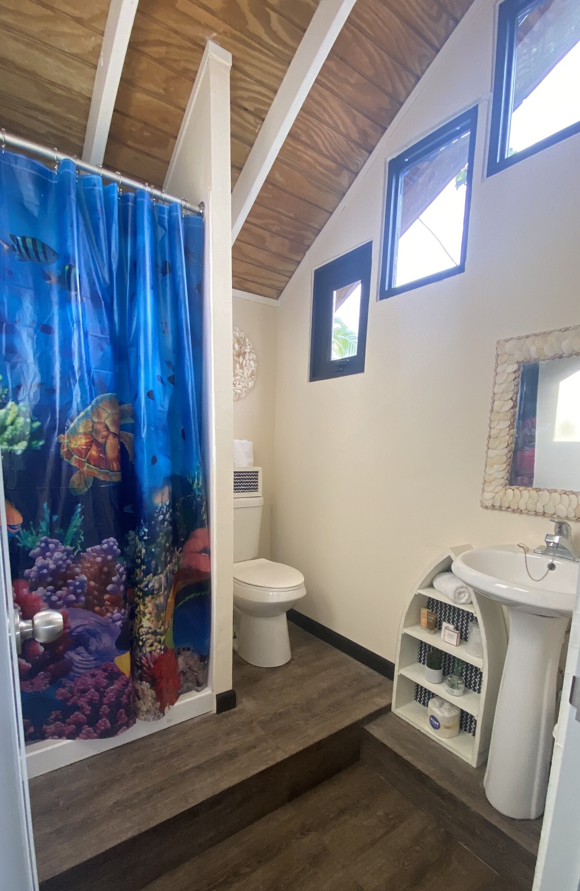 A bathroom with a shower curtain featuring an underwater scene, a toilet, a sink with a mirror, and a small shelf with toiletries.