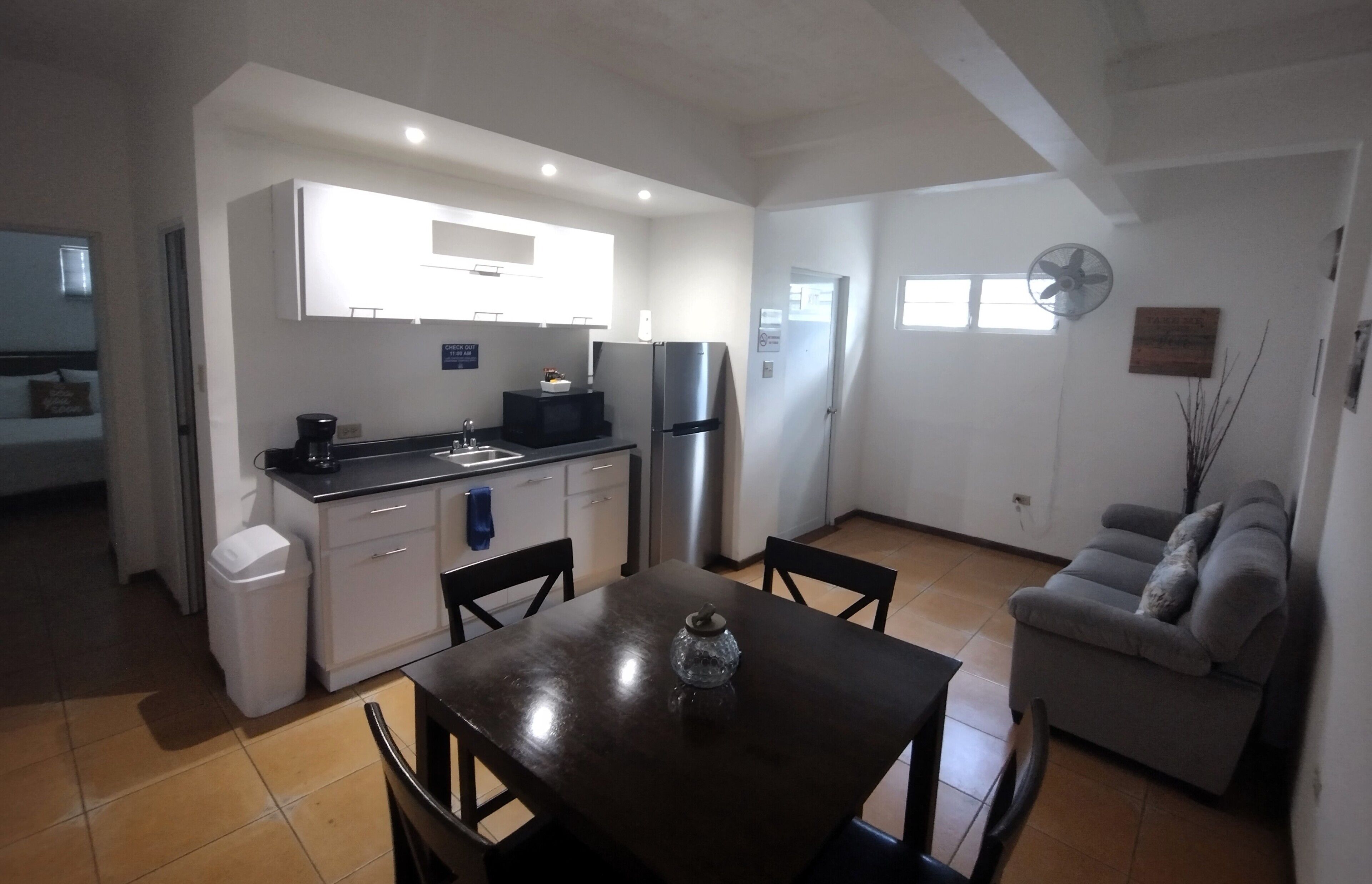 A compact kitchen with white cabinets, a black countertop, and a stainless steel refrigerator. There is a dining table with chairs in the foreground.