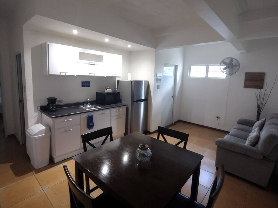 A compact kitchen with white cabinets, a black countertop, and a stainless steel refrigerator. There is a dining table with chairs in the foreground.